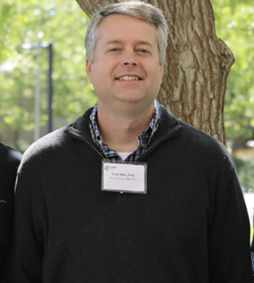 White man with grey hair wearing a black sweater over a blue, checkered, long-sleeved shirt, stands in front of a tree andsmiles at camera.